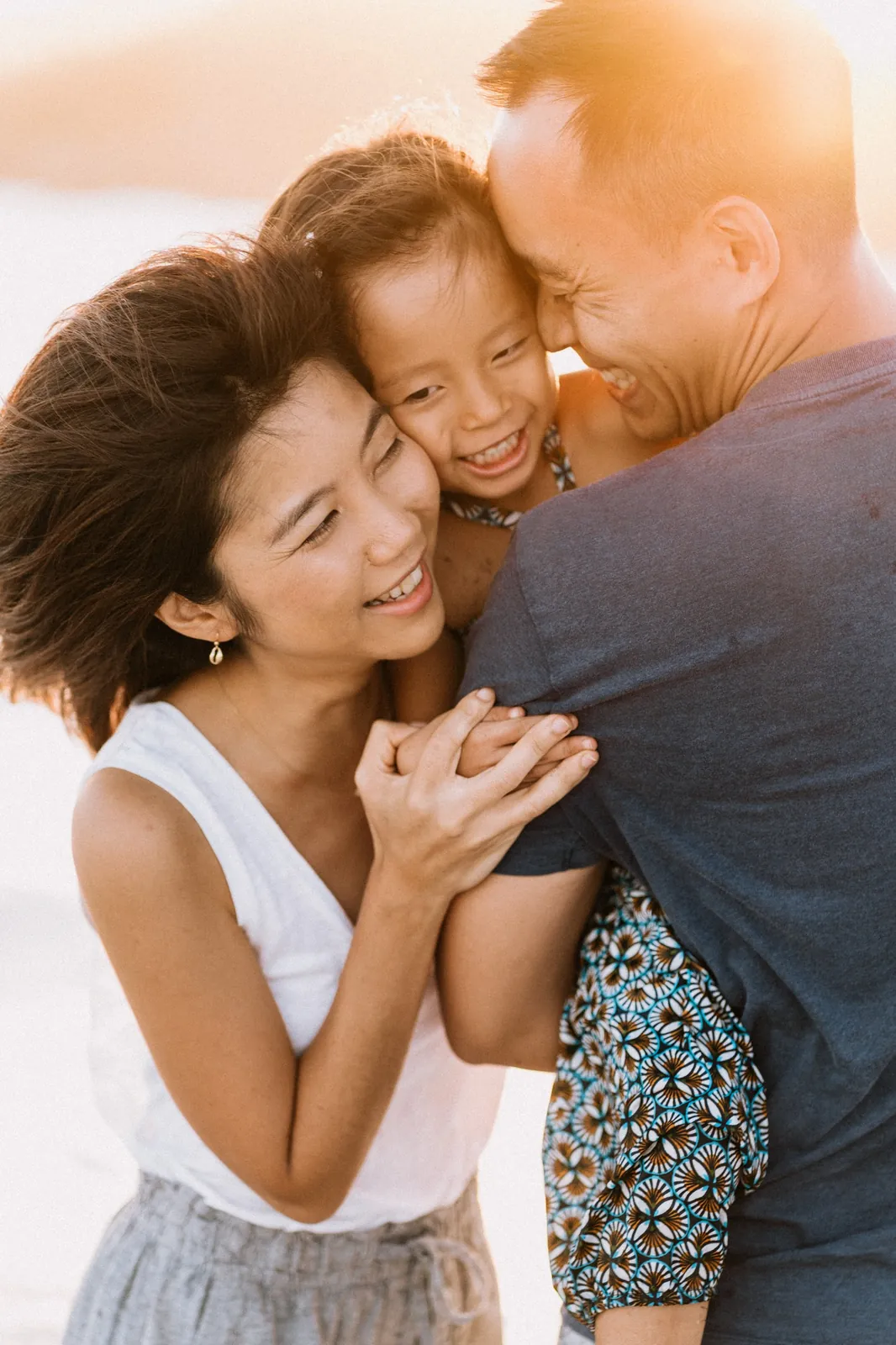 Au coucher du soleil, une petite fille rit aux éclats, serrée joue contre joue entre sa maman et son papa
