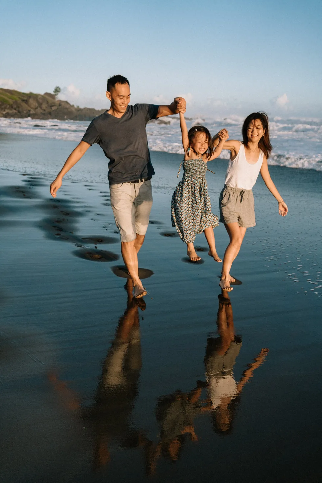 Sur une plage de sable noir, les parents soulèvent leur petite fille qui rit, les pieds en l'air, leurs reflets dans l'eau